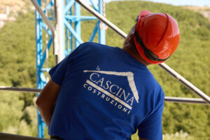 Inquadratura da dietro di un operaio edile con casco rosso e maglietta blu con il logo "La Cascina Costruzioni" su un ponteggio con vegetazione sullo sfondo