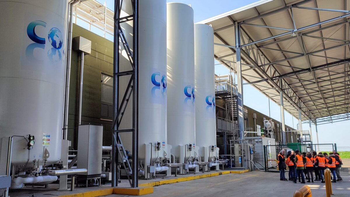 Una fila di alti serbatoi cilindrici bianchi con il simbolo della CO2 affiancati a una struttura industriale coperta, con un gruppo di visitatori in gilet ad alta visibilità sullo sfondo
