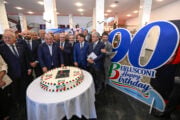 Un gruppo di uomini in abito scuro posa attorno a un tavolo con una grande torta decorata con i colori della bandiera italiana e il logo di Forza Italia, affiancata da un grande pannello blu e bianco con la scritta "90 Berlusconi Happy Birthday"