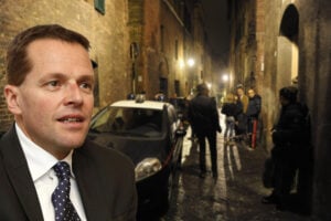 Primo piano di David Rossi in abito scuro sovrapposto all'immagine in notturna del vicolo di Siena dove è avvenuta la tragedia, con auto delle forze dell'ordine e persone presenti