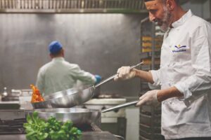 Chef in divisa Vivenda mentre cucina con una padella d'acciaio in un grande impianto di ristorazione
