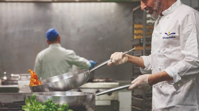 Chef in divisa Vivenda mentre cucina con una padella d'acciaio in un grande impianto di ristorazione
